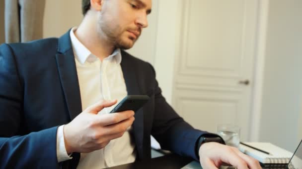 Jonge ernstige zakenman laptop gebruikt voor het werken in het bureau tijdens het chatten met vrienden op smartphone. — Stockvideo