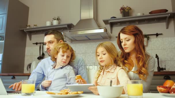 Famille prenant le petit déjeuner en cuisine. Petit garçon assis sur le père et la fille assis sur la mère, le père en utilisant un ordinateur portable et dauther en utilisant une tablette . — Video