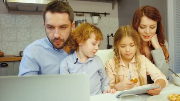 Οικογένεια με πατέρα που χρησιμοποιούν φορητό υπολογιστή, μητέρα, dauther και γιος χρησιμοποιώντας tablet χωροθέτηση σε φόντο κουζίνα. Εσωτερική. — Αρχείο Βίντεο