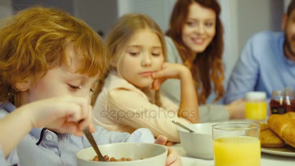 Família jovem caucasiana de pai, mãe, filha e filho pequeno tomando café da manhã na cozinha. Menino comendo flocos com leite . — Vídeo de Stock