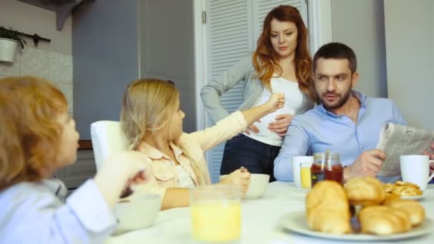 Niño, niña, padre y madre caucásicos en la cocina desayunando. Padre leyendo el periódico, madre de pie junto a él, dauther e hijo pequeño comiendo hojuelas con leche . — Vídeos de Stock