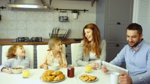 Familia joven caucásica en cocina. Padre leyendo el periódico, madre comiendo galletas, dauther y pequeño hijo comiendo hojuelas con leche . — Vídeo de stock