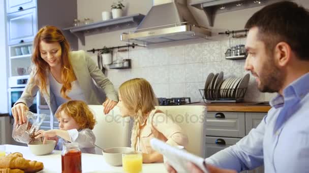 Kaukasische jongetje, meisje en vader aanbrengen op de tafel in de keuken. Vader krant lezen, moeder vullen vlokken in plaat van zoon, dauther vlokken met melk te eten. — Stockvideo