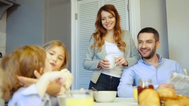 Kaukasische meisje eten van ontbijt en streelde haar broer, ouders kijken op hun en glimlachend op keuken achtergrond. — Stockvideo