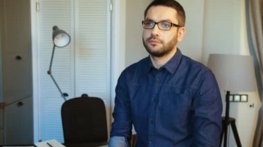 Gözlük modern ofiste siting ve gülümseyerek kameraya bakarak sakallı yakışıklı beyaz genç adam. Kapalı.