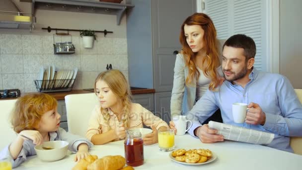 Kaukasische kleine jongen en meisje eten van de vlokken met de melk, vader en moeder drinken koffie op de achtergrond van de keuken. Indoor. — Stockvideo