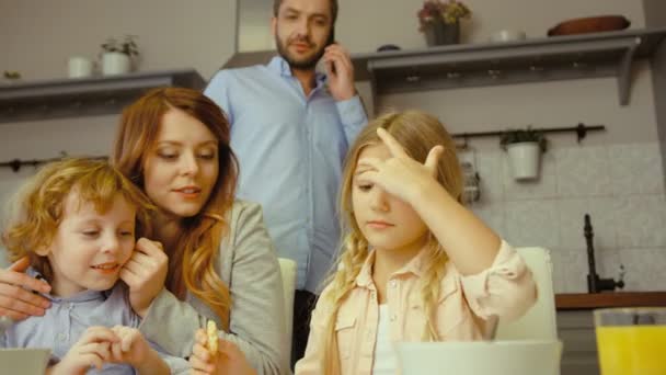 Caucasienne jolie femme avec ses enfants charmants regardant une tablette numérique tandis que son mari parle sur le téléphone intelligent dans la cuisine . — Video