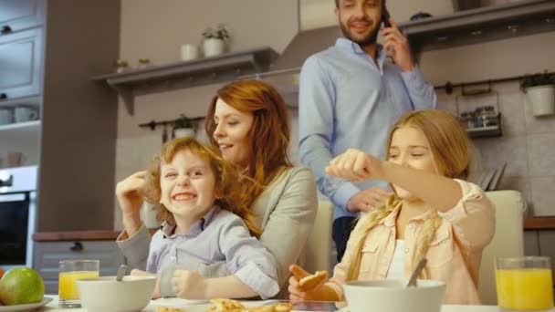 Donna caucasica con i suoi bellissimi figli che guardano un tablet digitale mentre suo marito parla al cellulare in cucina . — Video Stock