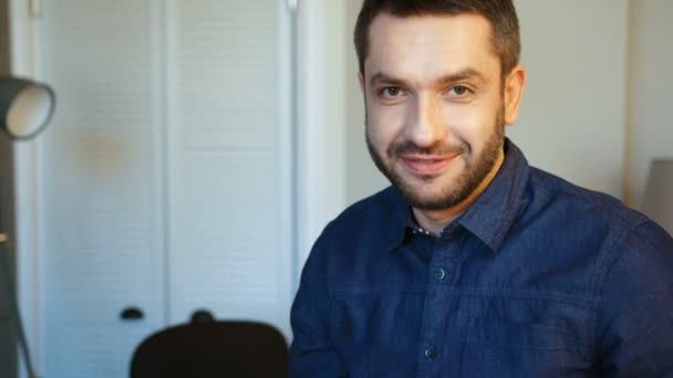 Retrato de hombre guapo caucásico con barba en camisa azul poniendo gafas, mirando a la cámara y sonriendo. De interior . — Vídeos de Stock