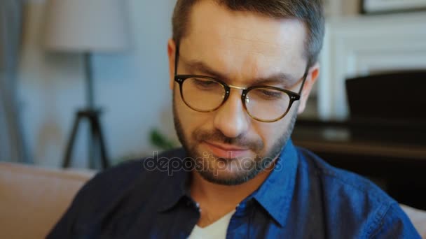 Портрет кавказского привлекательного мужчины в очках и с бородой в синей рубашке, читающего книгу на диване дома. В помещении . — стоковое видео
