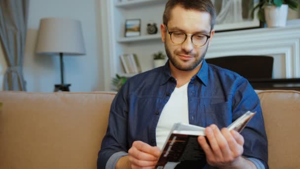 Jeune homme caucasien avec des lunettes et barbe en chemise bleue lecture livre assis sur le canapé à la maison. Intérieur . — Video