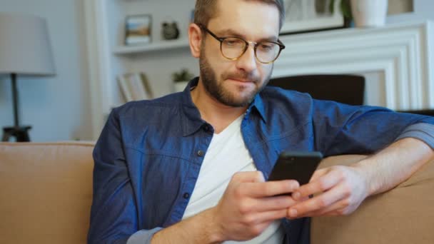 Ελκυστική Καυκάσιος άντρας με γυαλιά και τη γενειάδα χωροθέτηση στον καναπέ χρησιμοποιώντας το κινητό τηλέφωνο για την αναπαραγωγή παιχνιδιών στο σαλόνι. Εσωτερική. — Αρχείο Βίντεο