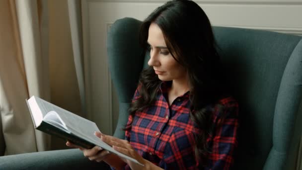 Mujer joven interesada leyendo libro mientras está sentado en una silla cómoda en la sala de estar. De cerca. Voltear páginas — Vídeos de Stock