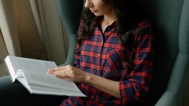 Jeune femme attrayante lisant livre intéressant tout en étant assis sur une chaise confortable dans le salon. Gros plan — Video