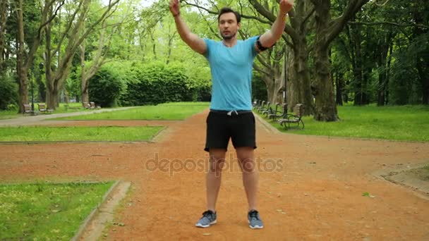 Unga stiliga fitness man gör stretching övningar med händerna i en vacker grön park innan du kör. Slow motion. — Stockvideo