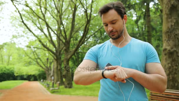 朝の公園でのスマートな腕時計と、実行する前にスマート フォンを使用して魅力的なハンサムなフィット男。クローズ アップ — ストック動画