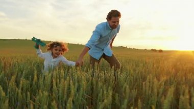 Küçük oğlu runing ahşap uçak oyuncak ile yeşil buğday alanı ile el ele tutuşarak ile genç baba. Uçmak gibi davranarak