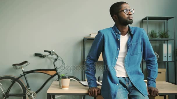 Retrato de un joven y atractivo hombre de negocios africano con gafas en camisa azul denim mirando a la cámara y sonriendo en el fondo de la oficina mientras se apoya en la mesa de la oficina . — Vídeos de Stock