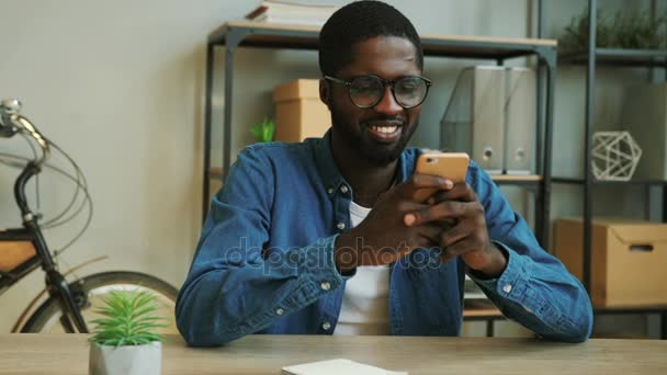 Portrait of smiling african business man in denim blue shirt using smart phone for chating with friends in the work time. Close up. — Stock Video