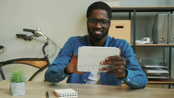 Retrato del hombre de negocios africano sonriente en camisa azul denim usando tableta para charlar con amigos en el descanso del lanch. De cerca. . — Vídeo de stock