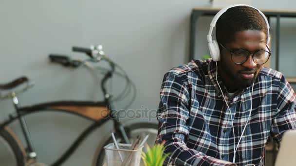 Retrato del joven empresario africano en glasess escuchando música en auriculares mientras escribe en el portátil en la oficina moderna . — Vídeo de stock