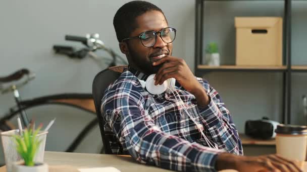 Retrato de un joven hombre de negocios africano en glasess y auriculares usando una computadora portátil para escribir el informe mientras está sentado a la mesa en la oficina moderna . — Vídeos de Stock