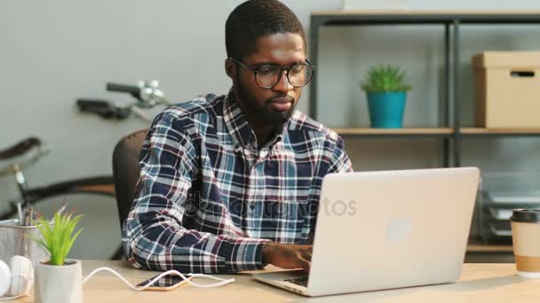 Porträt eines jungen afrikanischen Geschäftsmannes in lässigem Hemd und Brille, der den Bericht im modernen Büro am Laptop schreibt. — Stockvideo