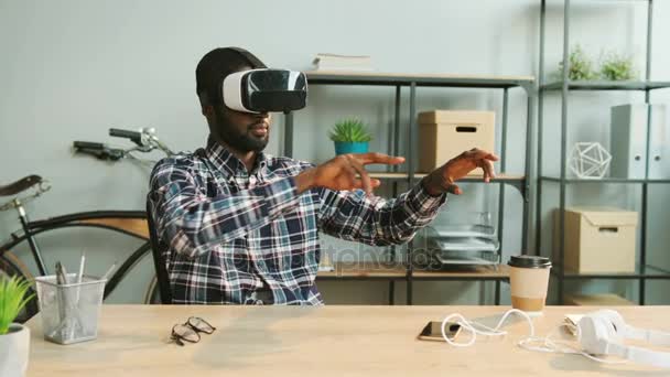 Zwarte Afrikaanse man in casual shirt met behulp van vr bril bij het moderne kantoor voor het bekijken van interessante video zittend aan tafel. — Stockvideo