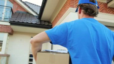 Paketi sahibine teslim etmek için eve kartonla yürüyen kurye portresini kapat. Arkadan bak..