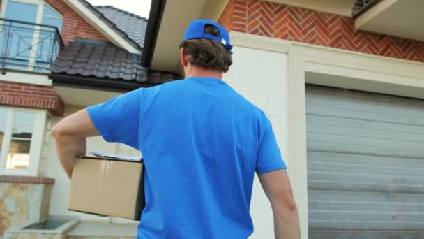 Correio andando com papelão para a casa para entregar o pacote ao proprietário. Vista pelas traseiras. Fechar tiro . — Vídeo de Stock