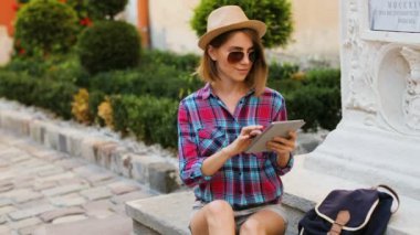 Şehir bankta dinlenirken tablet içinde yazarak şapka ve güneş gözlüklü genç beyaz kadın.