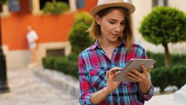 Joven mujer guapa en el sombrero usando la tableta mientras se relaja en el banco en la calle de la ciudad . — Vídeos de Stock