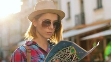 Genç çekici kadın şık şapka ve güneş gözlüğü güzel eski şehirde günbatımı sırasında seyahat ederken doğru yolu bulmak için harita kullanarak portresi.