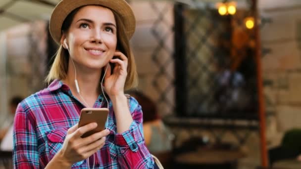 Mujer joven y hermosa en el sombrero de moda escuchando música en los auriculares con el teléfono inteligente y mirando a su alrededor en la ciudad vieja. Retrato plano . — Vídeo de stock