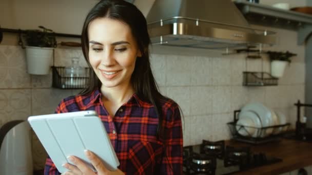 Mujer hermosa joven usando tableta mientras está de pie en la cocina. Mujer navegando por internet, leyendo la receta. De cerca. — Vídeos de Stock