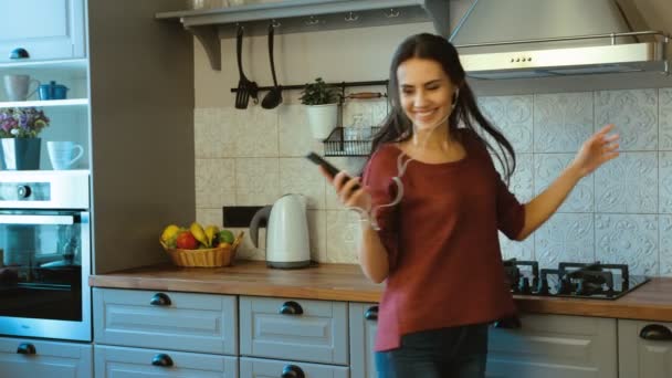 Mulher atraente feliz ouvir música com fones de ouvido em seu telefone inteligente e dançar na cozinha. Fechar — Vídeo de Stock