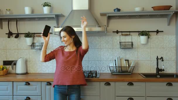 Hermosa mujer feliz escuchando música con auriculares en su teléfono inteligente y bailando en la cocina. Mujer sonriendo y bailando al ritmo. De cerca. — Vídeo de stock
