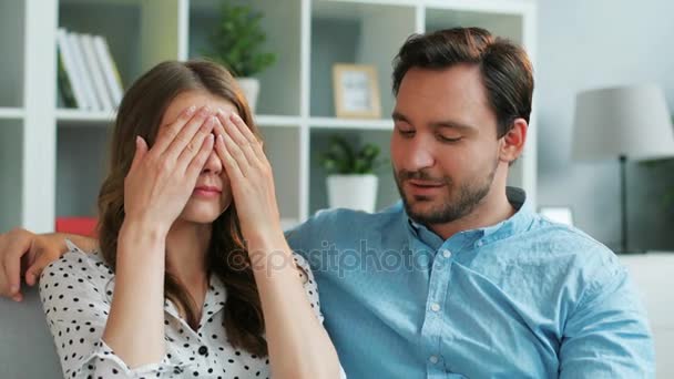 Homem atraente surpreendendo sua namorada emocional que fechando os olhos com um presente enquanto eles se sentam no sofá em casa . — Vídeo de Stock
