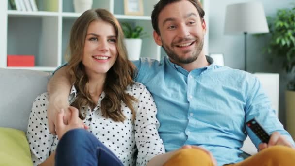 Porträt eines jungen verliebten emotionalen Paares, das zu Hause auf dem Sofa den Fernseher anschaut, sich umarmt und gemeinsam lacht. — Stockvideo