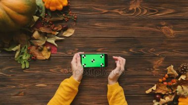 Kahverengi ahşap masa üzerine havadan görünümü sonbahar akçaağaç yaprakları ve kabak. Kaydırma, sürgülü, siyah akıllı telefon yatay pozisyonda yeşil ekran ile dokunarak kadın. Chroma anahtar. Hareket izleme