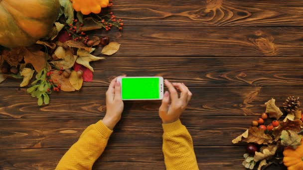 Teléfono inteligente blanco con pantalla verde en mesa de madera en posición horizontal. Vista superior de otoño con hojas de arce y calabaza. Mujer tocando en la pantalla táctil. Clave de croma — Vídeo de stock