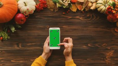 Kaydırma, dokunarak, ahşap masa üzerinde beyaz smartphone yeşil ekran yakınlaştırma kadın. Kabak üstünde belgili tanımlık tepe ile sonbahar kompozisyon. Üstten Görünüm. Chroma anahtar. Hareket izleme