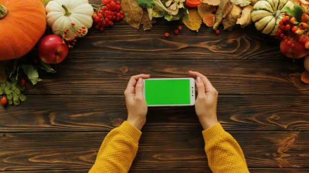 White smartphone with green screen in horizontal position on the wooden table with pumpkins. Top view. Female finger scrolling pages. Autumn. Chroma key — Stock Video