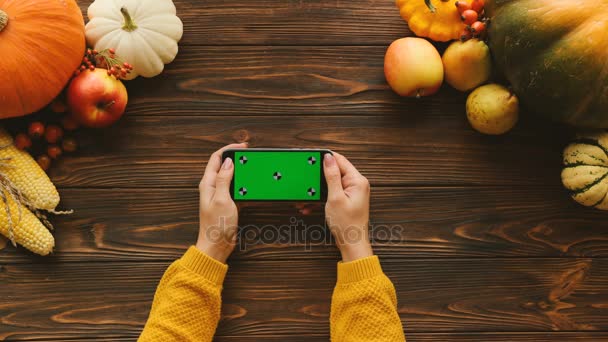 Осенний состав овощей и фруктов на деревянном столе. Верхний вид снимка женских рук, прокручивающихся, постукивающих по черному смартфону с зеленым экраном в горизонтальном положении. Хрома-ключ. Отслеживание — стоковое видео