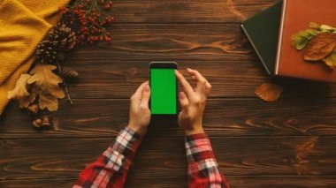 Kadın kaydırma dokunarak, ahşap masa üstünde yeşil perde ile siyah akıllı telefon dokunmatik ekran yakınlaştırma. Üstten Görünüm kitapları ile akçaağaç yaprakları, kazak. Dikey konum. Chroma anahtar