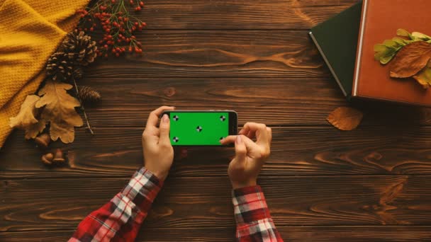 Vrouw over het scherm te onttrekken, touchscreen zoomen op zwarte smart phone met groen scherm op de houten tafel. Bovenaanzicht met boeken, esdoorn bladeren, trui. Horizontale positie. Chromakey. Volgen van beweging — Stockvideo