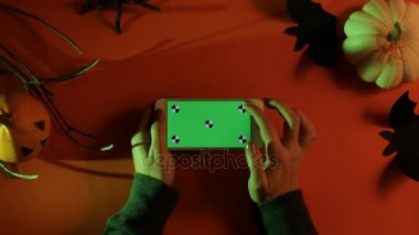 Cadılar Bayramı dekorasyonu ile turuncu zemin üzerine yeşil ekran ile beyaz akıllı telefon. Kabaklar, yarasalar, örümcek. Üstten Görünüm. Düz yatıyordu. Chroma anahtar. Hareket izleme