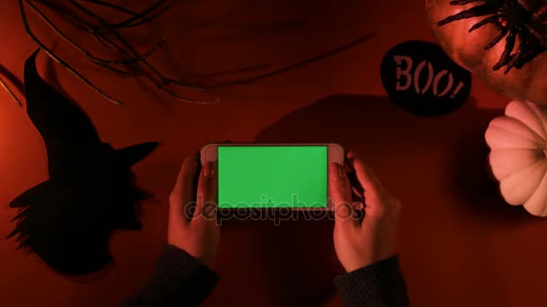 Vrouw met behulp van witte smartphone met groen scherm op oranje tafel met halloween decor. Bovenaanzicht. Chromakey — Stockvideo