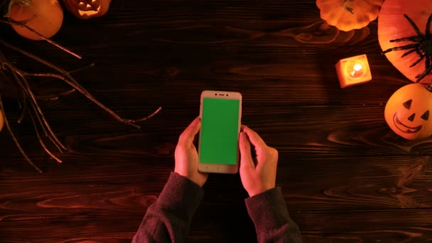 Mujer deslizando, tocando en el teléfono inteligente con pantalla verde. Vista superior de Halloween con calabazas de miedo en la mesa de madera. Clave de croma — Vídeos de Stock