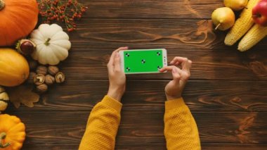Ahşap masa üzerinde yatay konumda yeşil ekran ile beyaz smartphone kullanan kadın. Masada sonbahar üstten görünüm meyve ve sebze ile. Chroma anahtar. Hareket izleme